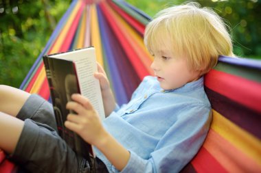 Küçük sevimli sarışın beyaz çocuk kitap okuyor ve arka bahçede ya da dışarıda renkli hamakla eğleniyor. Çocuklar için yaz tatili. Hamakta sallanan ve rahatlatan çocuk.