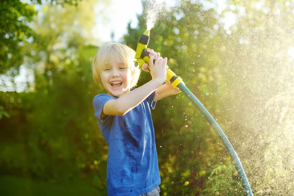 面白い小さな男の子は植物を散水し 日当たりの良い裏庭でスプリンクラーで庭のホースで遊んでいます 水のスプレーで楽しんでいる未就学児 子供のための夏の屋外活動 — ストック写真