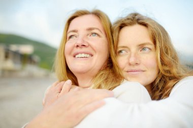 Kızıl saçlı kıdemli anne ve güzel yetişkin kızı kucaklaşıyorlar. Bir anne ve yetişkin çocuğunun mutlu bir buluşması. Yetişkin çocuklar ve yaşlı ebeveynler arasındaki aile ilişkileri