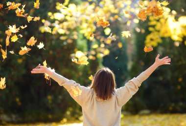 Young woman is having fun while walking through the forest on a sunny autumn day. Girl plays with maple leaves and throws them up. Fallen leaves rustle.