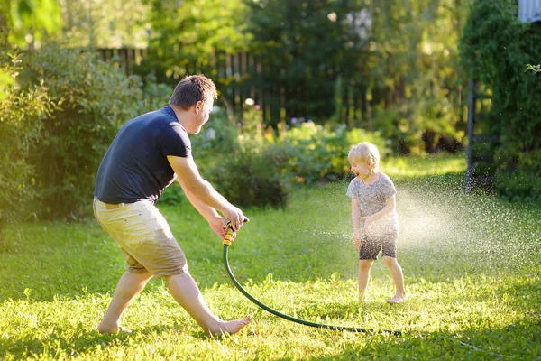 彼の父は 日当たりの良い家の裏庭の庭のホースで遊んで面白い小さな男の子 未就学児の子は 水のスプレーを楽しんでします 子供のための夏の屋外活動 — ストック写真