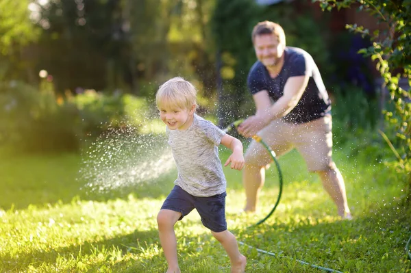 有趣的小男孩与他的父亲在阳光明媚的后院玩花园软管 学龄前儿童享受水的喷水的乐趣 儿童夏季户外活动 — 图库照片