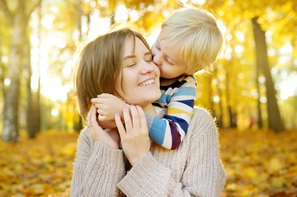小さな男の子は彼の若い母親を抱擁し 晴れた秋の公園で散歩中に彼女にキスしています ママと子供の間の愛と感情 — ストック写真