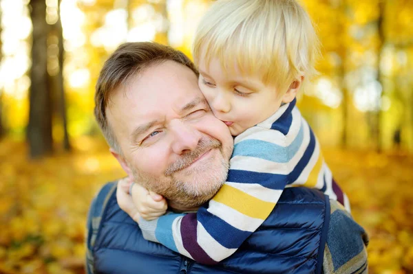 小さな男の子は彼の成熟した父親を受け入れ 晴れた秋の公園を散歩中にキスします 父親と子どもの間の愛情と感情 — ストック写真