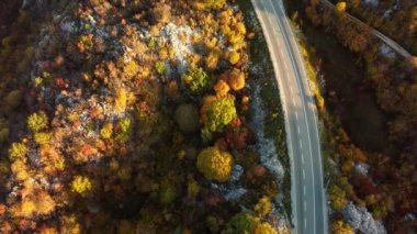 Panoramik sonbahar sonlarında nefes kesici hava aracının görüntüsü. Tepelerin üst hava manzarası ve arka planda yüksek dağ zirveleri olan günbatımı ışıklarıyla yıkanmış bir hız yolu. Doğadaki mevsimlerin güzelliği