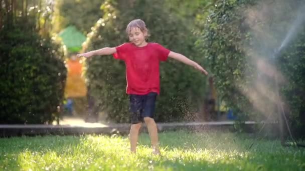 日当たりの良い裏庭で庭のスプリンクラーで遊んで面白い男の子のスローモーションビデオ 小学生は笑って ジャンプし 水のスプレーで楽しんでいます 子供のための夏の屋外活動 — ストック動画