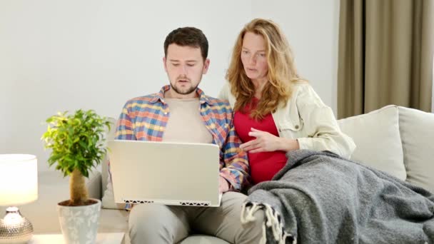一对夫妇在家里坐在沙发上 用他们的现代笔记本电脑在网上选择一些东西 一个年轻的家庭在等着孩子 怀孕妇女与丈夫讨论大宗购买或旅行 — 图库视频影像