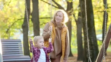 Bir anne oğlunu sonbahar parkında salıncakta sallıyor. Güzel bir kadın, sevimli bebeğini oyun alanında eğlendirir. Çocuklu aileler için geleneksel aktif mevsimlik eğlence
