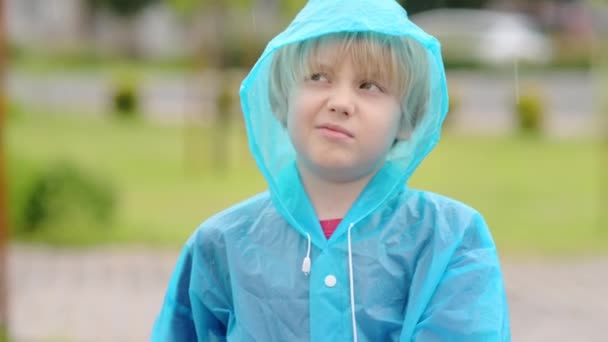 一个不满的孩子在雨中的画像 穿着蓝色雨衣的不满男孩对走路时的倾盆大雨不满意 年轻人对恶劣的天气已经厌倦了 — 图库视频影像