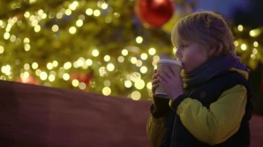 Tatlı çocuk sıcak çikolata içiyor ya da süslenmiş sokak Xmas ağacının yanında sıcak meyve suyu içiyor. Mutlu bir çocuk Noel arifesini kasaba meydanında kutluyor. Kış tatili gelenekleri