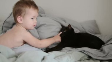 Sevimli çocuk, ebeveyn yatağında şekerleme yaptıktan sonra uyandı. Neşeli bebek ebeveyn yatağında siyah kediyle oynuyor. Çocuklarla evcil hayvanlar arasındaki ilişki