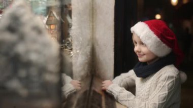Noel Baba şapkası takan sevimli çocuk kış akşamları mağaza vitrininde Noel süslerine hayran. Geleneksel Noel pazarında Xmas oyuncaklarına ve aksesuarlarına bakan turistler