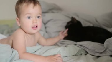 Sevimli çocuk, ebeveyn yatağında şekerleme yaptıktan sonra uyandı. Neşeli bebek ebeveyn yatağında siyah kediyle oynuyor. Çocuklarla evcil hayvanlar arasındaki ilişki