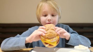 Fast food restoranında büyük hamburger yiyen tatlı sarışın çocuk. Çocuklar için sağlıksız bir yemek. Abur cubur. Aşırı kilolu çocuk.