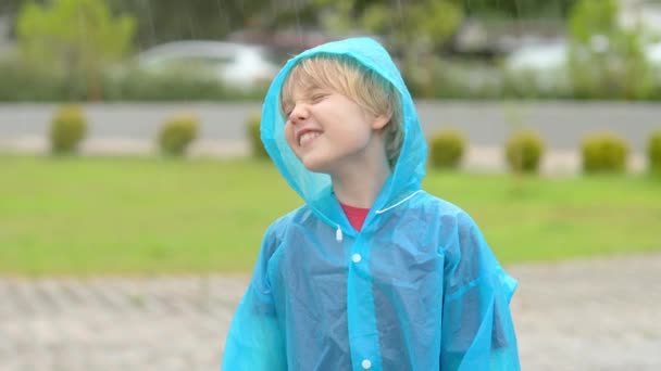 雨を降らせている幸せな子供の肖像 青いレインコートに乗った楽しい少年が 歩きながら激しい雨の中でジャンプして踊ります 子供は雨天で走って遊ぶのが大好きです — ストック動画