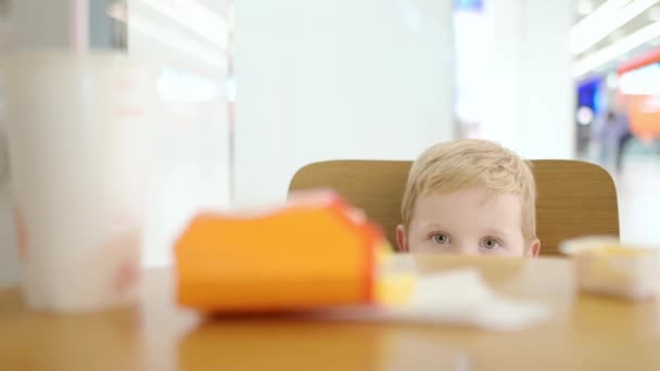 一个可爱的金发小孩躲在桌子后面 小孩子很害羞 不和别人接触 在快餐店里胆小害羞的婴儿 — 图库视频影像