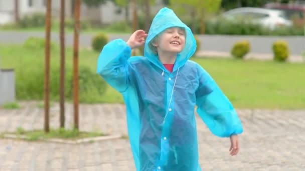 雨を降らせている幸せな子供の肖像 青いレインコートに乗った楽しい少年が 歩きながら激しい雨の中でジャンプして踊ります 子供は雨天で走って遊ぶのが大好きです — ストック動画