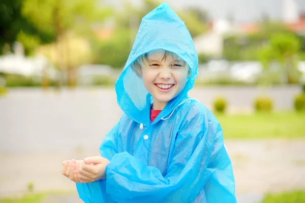 A joyful boy in a blue raincoat tries to catch raindrops in his palms while walking on a rainy day. Children love to run and play in rainy weather