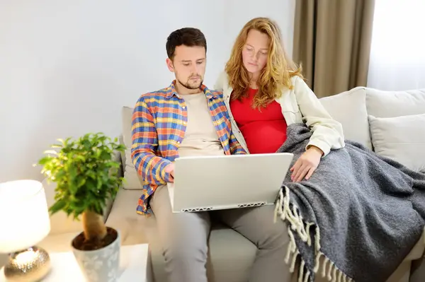 一对夫妇在家里坐在沙发上 用他们的现代笔记本电脑在网上选择一些东西 一个年轻的家庭在等着孩子 怀孕妇女与丈夫讨论大宗购买或旅行 图库图片