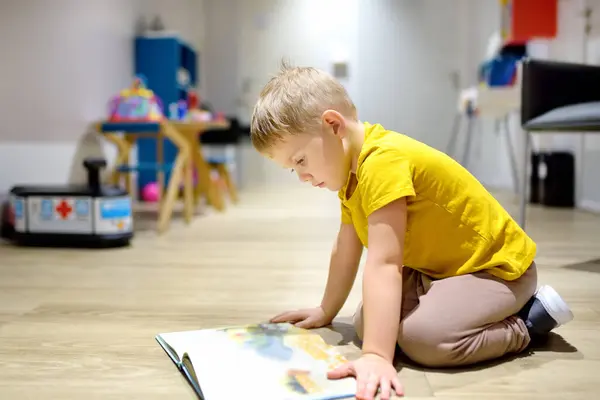 可爱的小男孩正在幼儿园的地板上看书 这孩子在幼儿园玩得很开心 图库图片