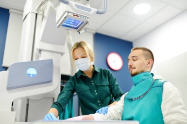 Modern klinikteki röntgen odasında genç bir adamın elinin röntgenini çekecek olan kadın radyolog. Hasta kurşun önlük takıyor. İnsanların tıbbi muayenesi
