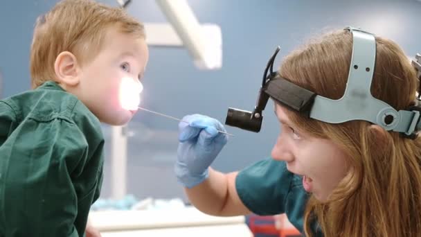 母親と一緒にいるかわいい幼児の少年が小児科医の診察を受けています 医師の耳鼻血管科医は 検査スパチュラを持つ小さな患者の喉を調べています ロイヤリティフリーストック映像