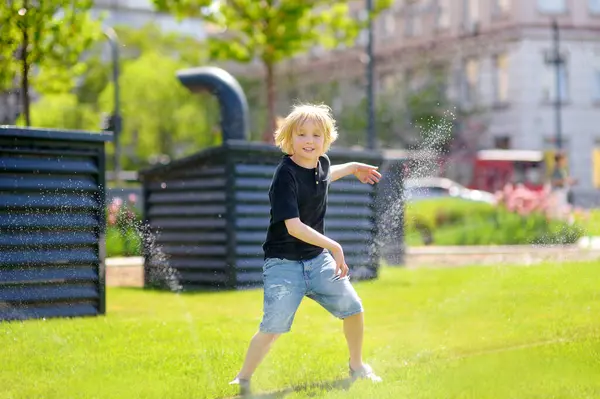 Divertente Ragazzino Che Gioca Con Irrigatore Nel Soleggiato Parco Cittadino Foto Stock