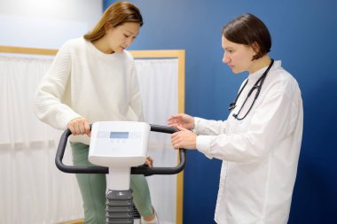 Female doctor cardiologist making bicycle electrocardiogram for examination cardiovascular system of patient at medical office of clinic. Young woman training on bike simulator. Exercise bike. clipart