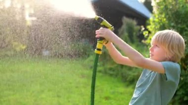 Komik çocuk serin su döküp sıcak yaz günlerinde bahçe hortumuyla güneşli arka bahçede fıskiyeyle oynuyor. Mutlu çocuk su sıçramalarıyla eğleniyor. Çocuklar için yaz açık hava aktivitesi.