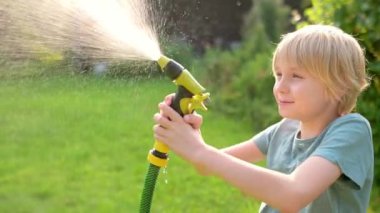 Komik çocuk serin su döküp sıcak yaz günlerinde bahçe hortumuyla güneşli arka bahçede fıskiyeyle oynuyor. Mutlu çocuk su sıçramalarıyla eğleniyor. Çocuklar için yaz açık hava aktivitesi.