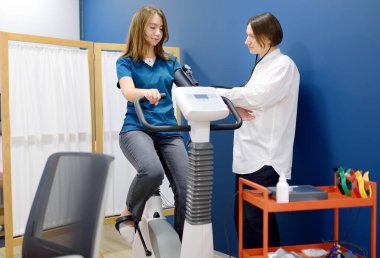 Kadın doktor kardiyolog, klinikteki kardiyovasküler sistemi incelemek için bisiklet egzersizi sırasında hastanın üzerindeki baskıyı ölçüyor. Bisiklet simülatöründe eğitim gören genç bir kadın. Egzersiz bisikleti