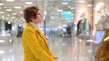 Alışveriş merkezinde alışveriş yaparken yaşlı bir kadının portresi. Güzel kızıl saçlı son sınıf öğrencisi vitrindeki mankenlerin üzerindeki şık kıyafetlere bakıyor.