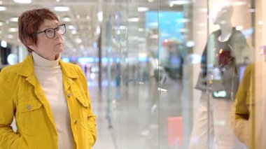 Alışveriş merkezinde alışveriş yaparken yaşlı bir kadının portresi. Kızıl saçlı son sınıf öğrencisi, şık ve ucuz kıyafetleri titizlikle seçerken vitrindeki mankenlerle ilgili yeni modellere bakıyor.