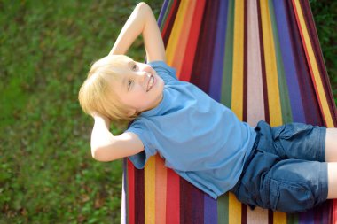 Sevimli sarışın beyaz çocuk arka bahçede ya da açık havada çok renkli hamakla eğleniyor. Çocuklar için açık havada aktif yaz tatili. Çocuk hamakta rahatlıyor ve sallanıyor. Görünümün üstünde