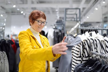 Alışveriş merkezinde alışveriş yaparken yaşlı bir kadının portresi. Kızıl saçlı son sınıf öğrencisi, şık ve ucuz kıyafetleri seçerken askılarda yeni modellere bakıyor.