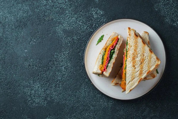 Three homemade sandwiches with sausage, cheese and arugula on a dark concrete background. The concept of a quick meal or snack at work or school. Top view with copy space