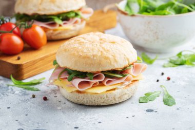 İki ev yapımı sandviç, jambon, peynir ve roka ile beton bir masada. Hafif arka planda iki hamburger..