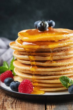 Close-up delicious pancakes, with fresh blueberries, raspberry and maple syrup or honey on a dark background. Sweet maple syrup flows from a stack of pancake