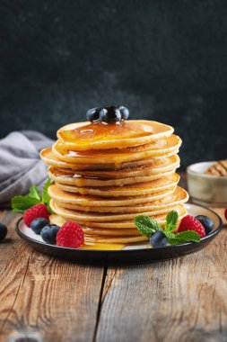 Close-up delicious pancakes, with fresh blueberries, raspberry and maple syrup or honey on a dark background. Sweet maple syrup flows from a stack of pancake