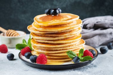 Close-up delicious pancakes, with fresh blueberries, raspberry and maple syrup or honey on a dark background. Sweet maple syrup flows from a stack of pancake