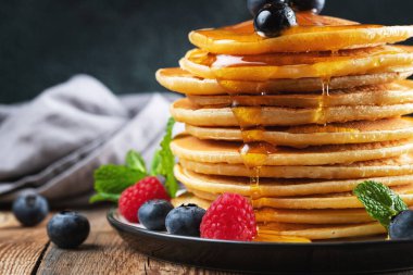 Close-up delicious pancakes, with fresh blueberries, raspberry and maple syrup or honey on a dark background. Sweet maple syrup flows from a stack of pancake