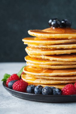 Close-up delicious pancakes, with fresh blueberries, raspberry and maple syrup or honey on a dark background. With copy space. Sweet maple syrup flows from a stack of pancake