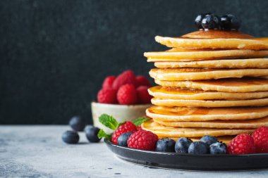 Close-up delicious pancakes, with fresh blueberries, raspberry and maple syrup or honey on a dark background. With copy space. Sweet maple syrup flows from a stack of pancake