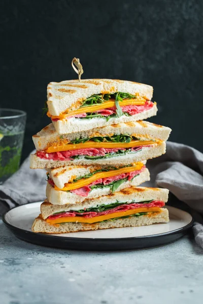 Big homemade sandwich with sausage, cheese and arugula on a dark concrete background. The concept of a quick meal or snack at work or school.