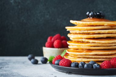 Close-up delicious pancakes, with fresh blueberries, raspberry and maple syrup or honey on a dark background. With copy space. Sweet maple syrup flows from a stack of pancake