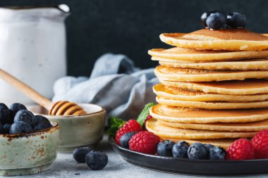 Close-up delicious pancakes, with fresh blueberries, raspberry and maple syrup or honey on a dark background. With copy space. Sweet maple syrup flows from a stack of pancake