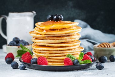 Close-up delicious pancakes, with fresh blueberries, raspberry and maple syrup or honey on a dark background. Sweet maple syrup flows from a stack of pancake