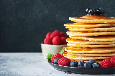 Close-up delicious pancakes, with fresh blueberries, raspberry and maple syrup or honey on a dark background. With copy space. Sweet maple syrup flows from a stack of pancake