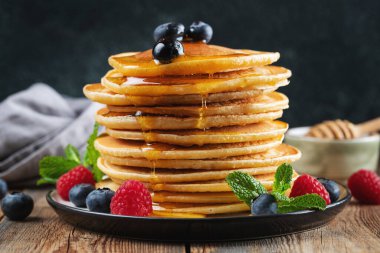 Close-up delicious pancakes, with fresh blueberries, raspberry and maple syrup or honey on a dark background. Sweet maple syrup flows from a stack of pancake
