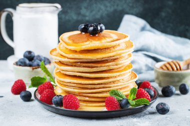 Close-up delicious pancakes, with fresh blueberries, raspberry and maple syrup or honey on a dark background. Sweet maple syrup flows from a stack of pancake